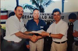 Fotografía donde destaca Victor Herrera estrechando su mano con una persona y en medio de ellos e...
