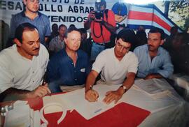 Fotografía de cuatro hombres, entre ellos Victor Herrera firmando un documento, Miguel Angel Rodr...