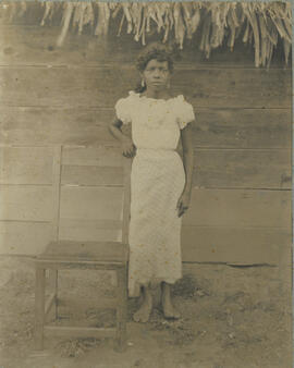 Álbum que contiene Fotografía de una mujer posando junto a una silla de madera, con un vestido bl...
