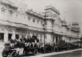 Fotografía con la imagen del desfile de carros y personal del cuerpo de bomberos, frente al edifi...